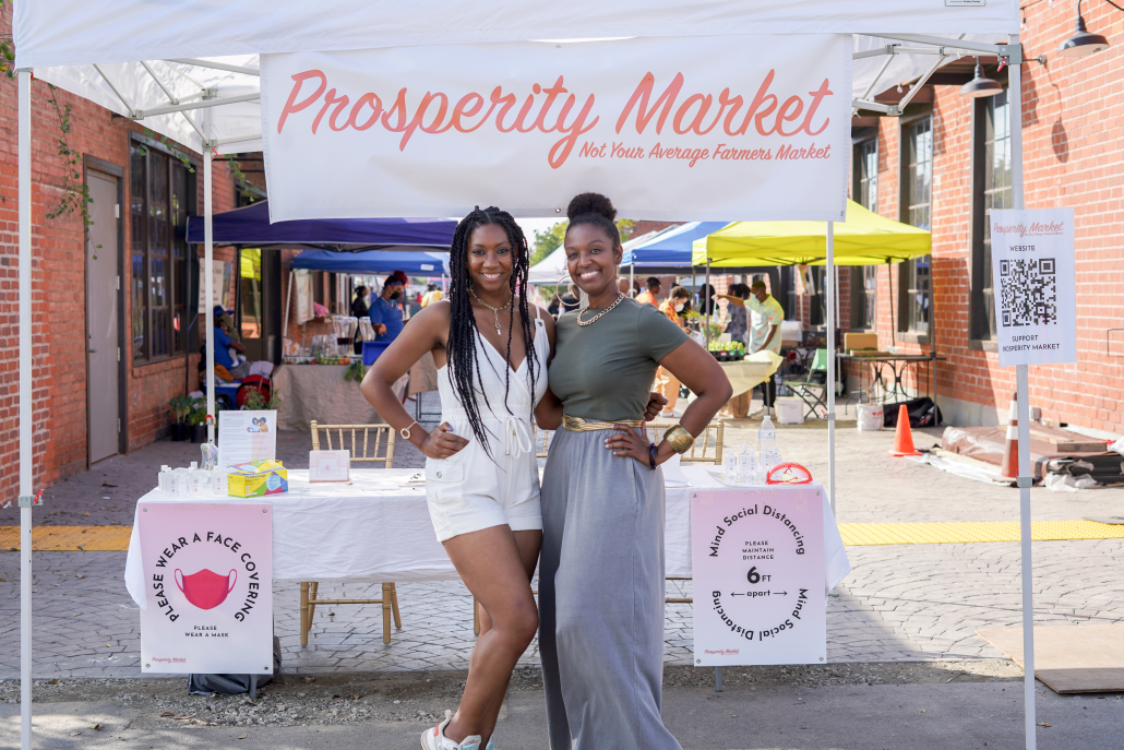Two women stand with arms around each others' waists, under banner with words "prosperity market"