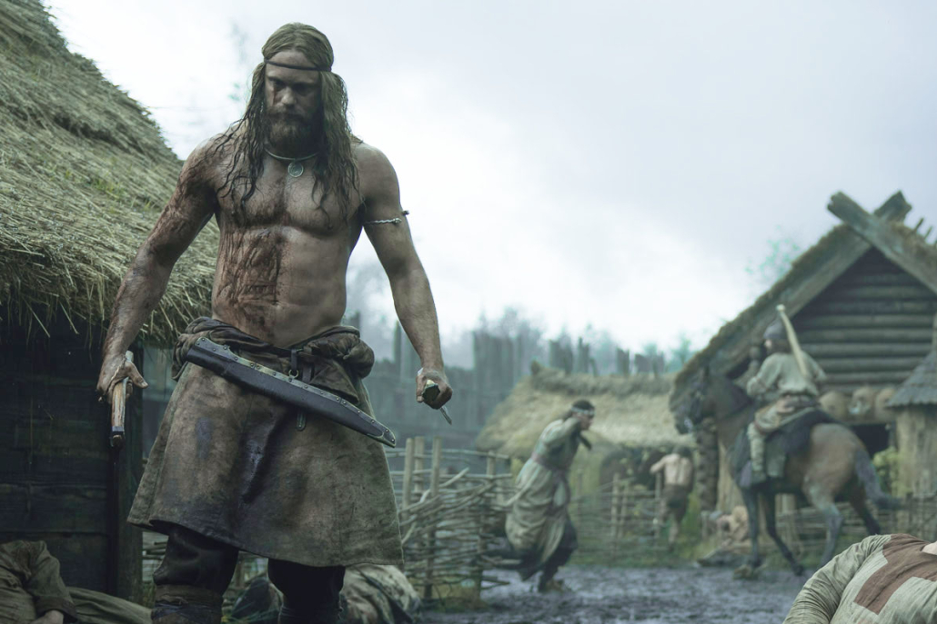 Alexander Skarsgård in the Northman as a Viking. He is standing with weapons in his hand in the middle of a village. He is also bloody. 