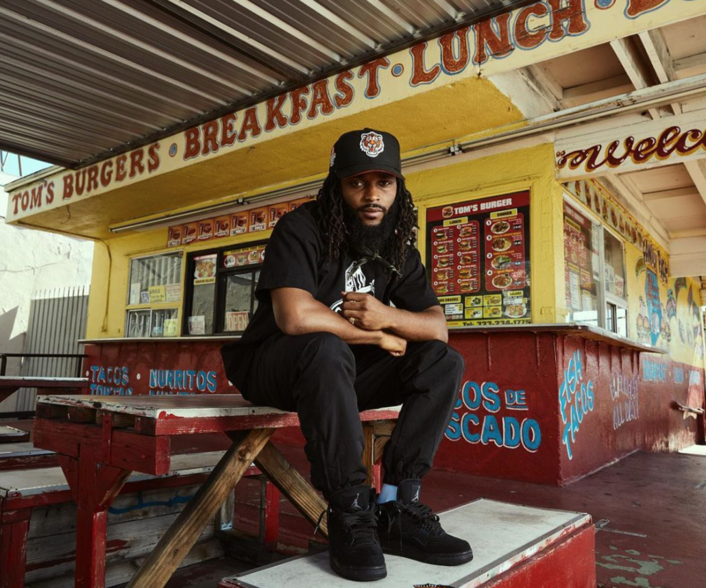 Sham1016 sits on a picnic table outside a South Central L.A. restaurant, Tom's Burger.