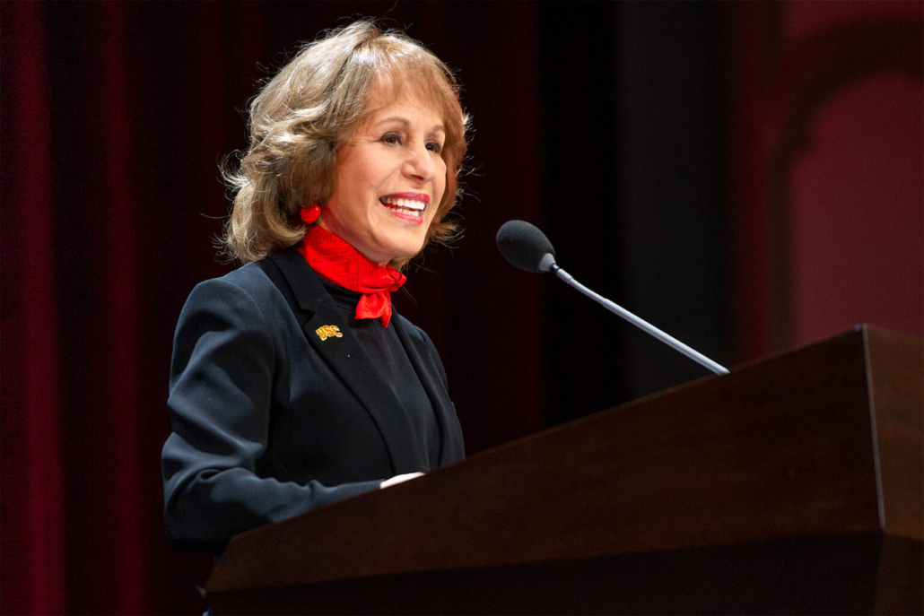 Carol Folt stands at a podium with a microphone. 