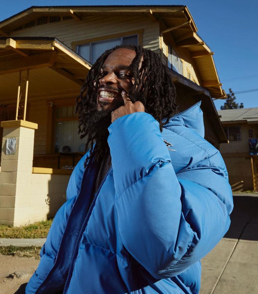 Rapper Sham1016 smiles at the camera, showing off a gold tooth, in front of a house in South Central.