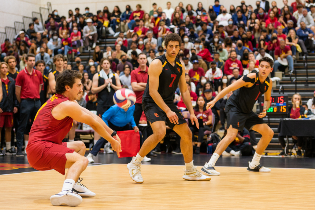 USC men's volleyball vs UCLA team. Libero squatted to bumps the ball, number 7 and number 1 supporting him. Blurry crowds in the back watching. 