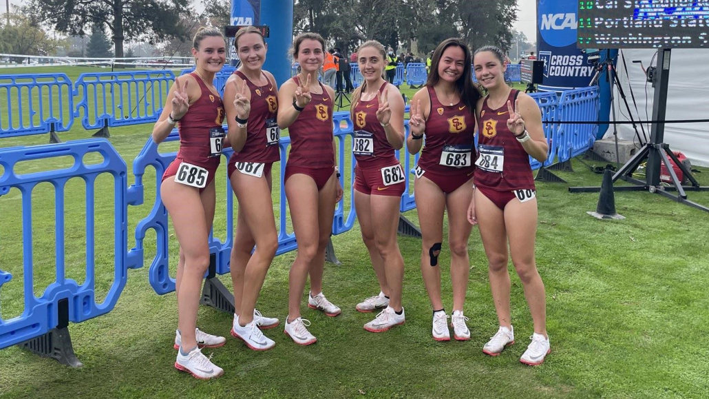 Image of six people in USC cross country uniforms holding up a piece sign. 