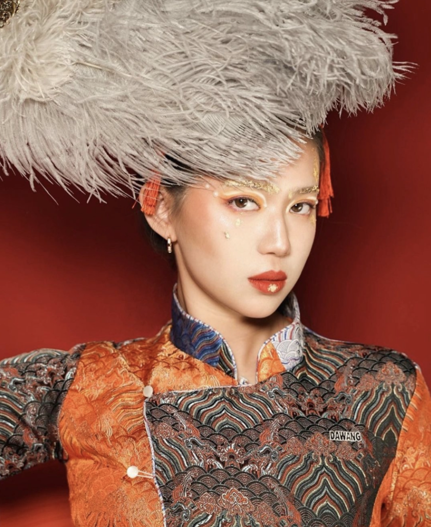 A model fanning herself with a large grey feather poses in a patchwork DAWANG shirt against a red background.