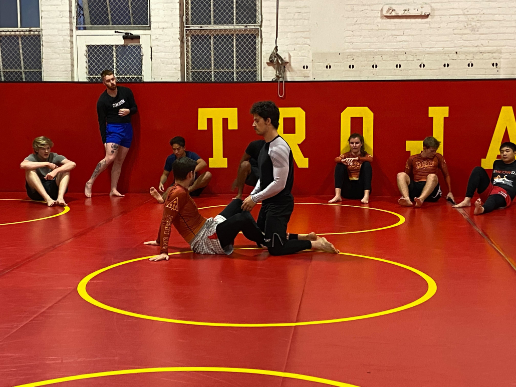 A fight club meeting. A guy pins another guy down on a red mat. Others sit around and watch.