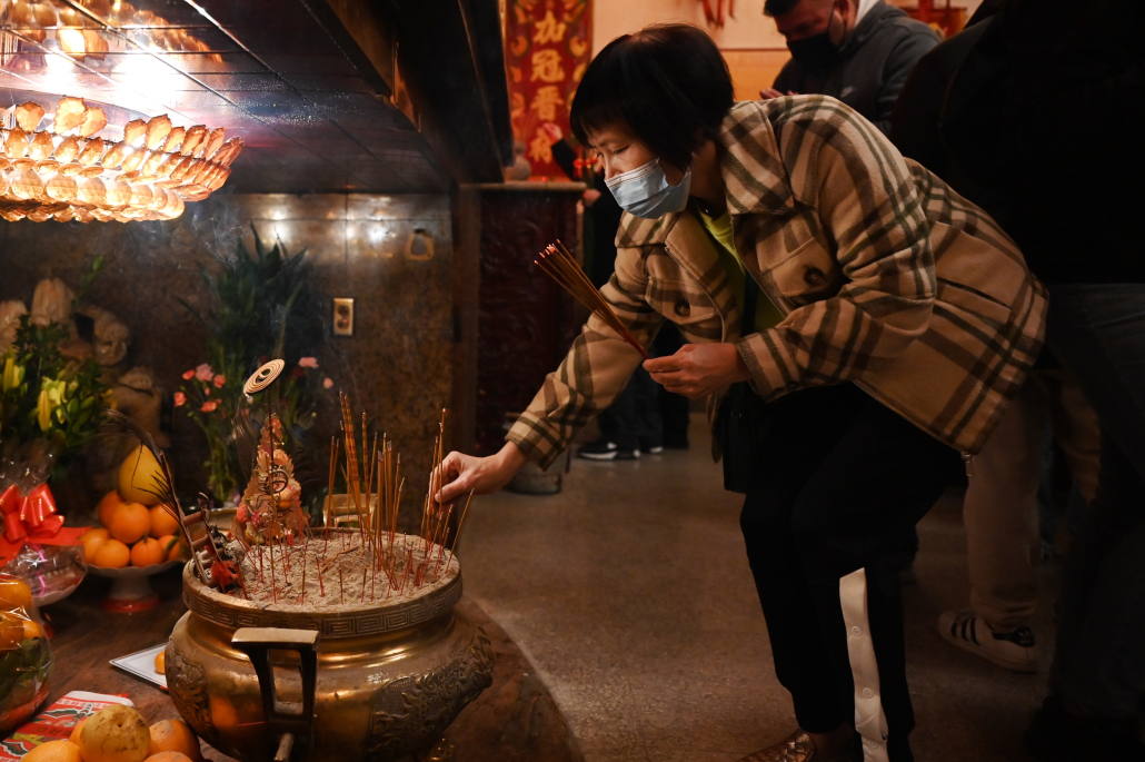 A masked woman reaches down to place a stick of incense onto an altar.