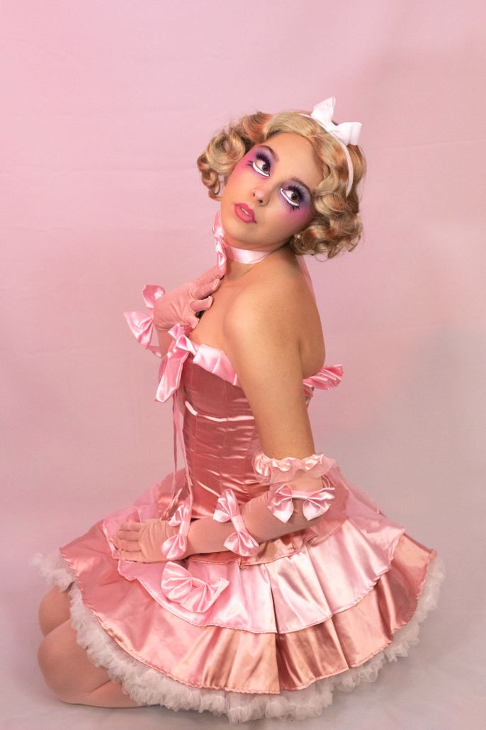 Daisy Darling kneels in a pink dress made of many bows. 