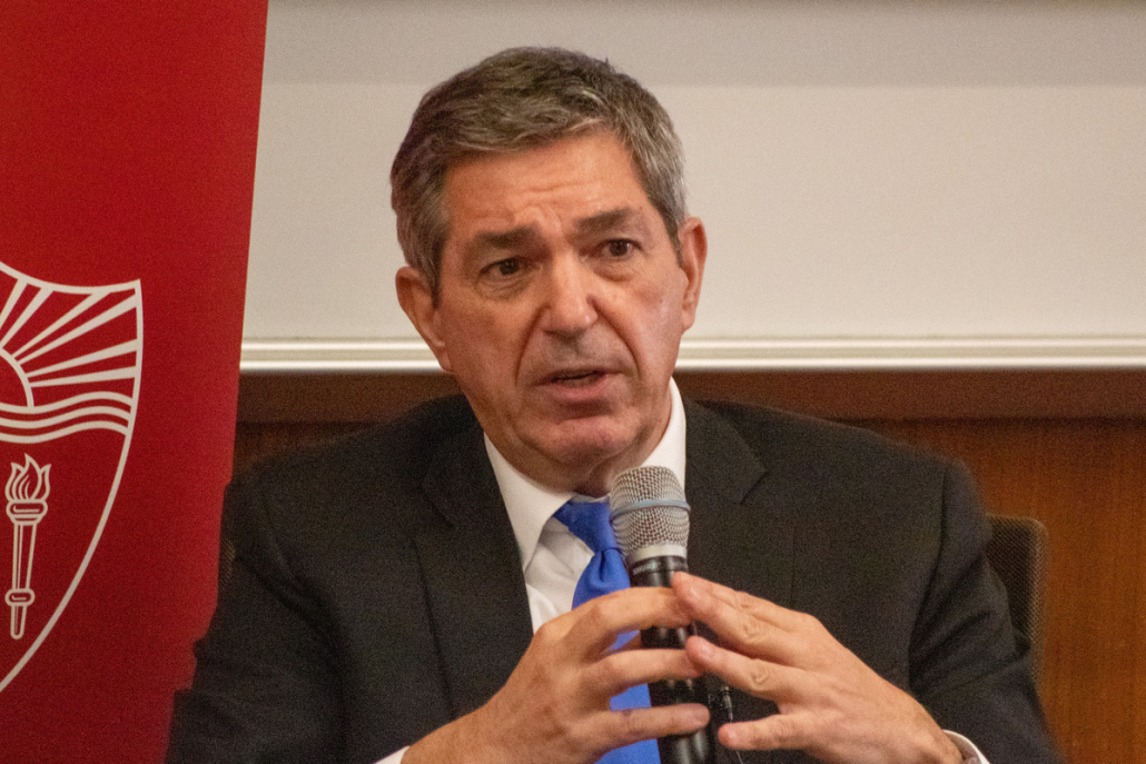 Stavros Lambrinidis sits with a microphone in his hands talking to the audience. He is wearing a black suit, a white button-down shirt and a bright blue tie.