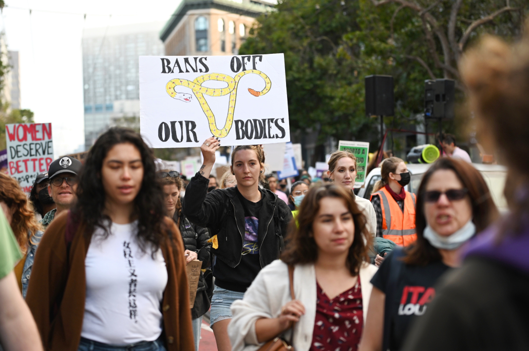 San Francisco protest after the overturn of Roe. Wade on June 24. A person wearing a black jacket and denim shorts holds up a sign reading, "Bans off our bodies." The sign also depicts a yellow snake coiled in the shape of an uterus.