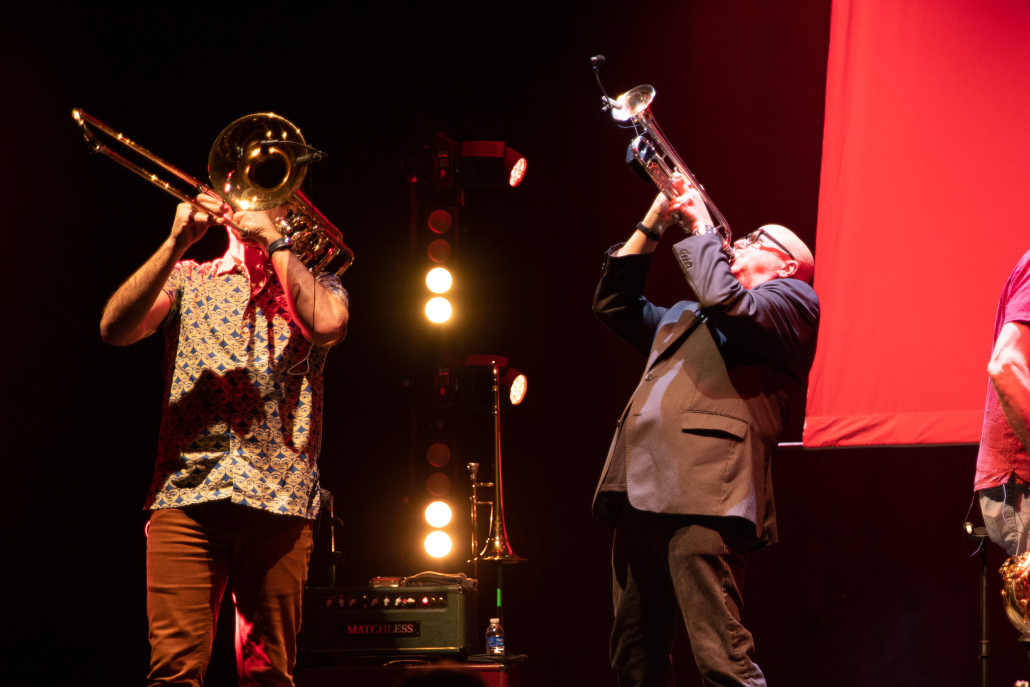 A photo of two members of They Might Be Giants playing trumpets on stage. 