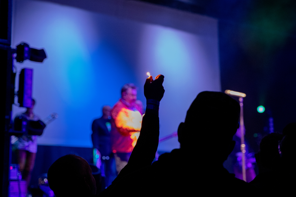 A photo of a person holding up a lighter in a crowd while They Might Be Giants performs on stage.