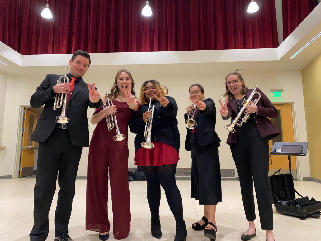 Photo of USC Trumpet Quintet holding up the "Fight on" sign while holding trumpets.