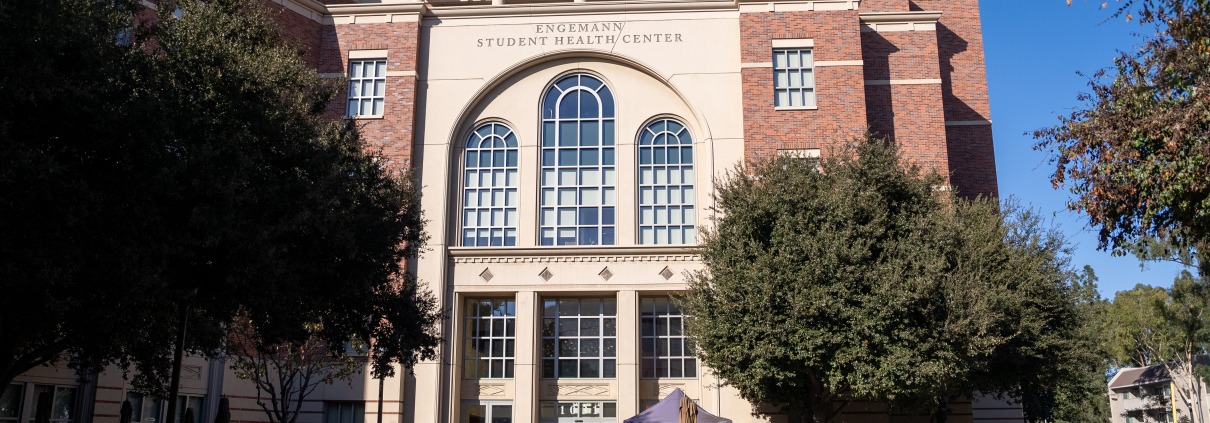 The entrance of the Engemann Student Health Center on a sunny day. The building is red brick with three columns of windows above the doors which rise up vertically before curving in a semicircle.