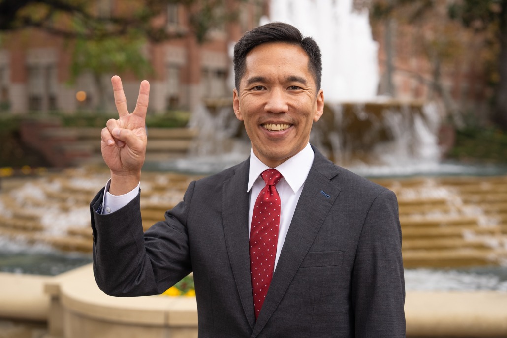 Beong-Soo Kim stands in front of the ShumWay fountain in a black suit and a red tie. He holds up a v for victory.