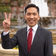 Beong-Soo Kim stands in front of the ShumWay fountain in a black suit and a red tie. He holds up a v for victory.