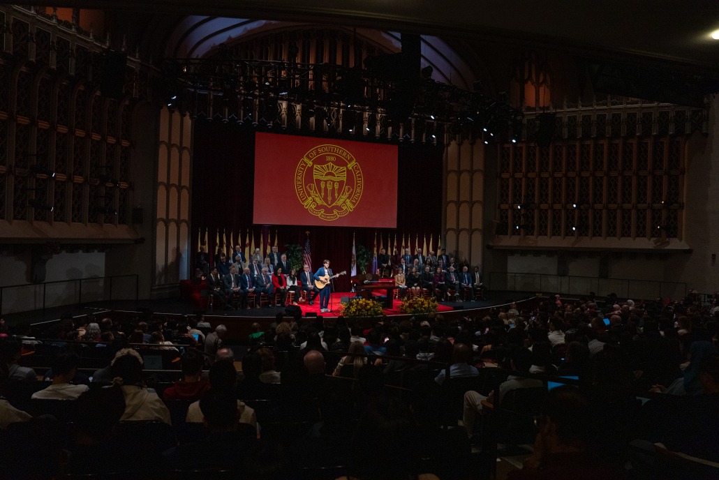 A large screen with the USC emblem acts as the background for a student singing and playing the guitar in front of hundreds inside Bovard Auditorium.