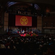 A large screen with the USC emblem acts as the background for a student singing and playing the guitar in front of hundreds inside Bovard Auditorium.