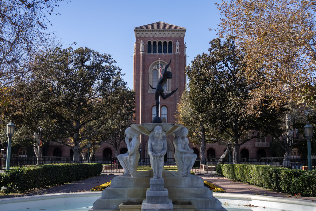 A statue of three hunched-over figures in Alumni Park in the foreground, with Bovard Auditorium in the background.