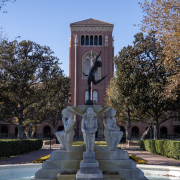 A statue of three hunched-over figures in Alumni Park in the foreground, with Bovard Auditorium in the background.