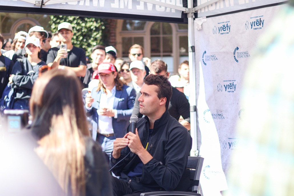 Charlie Kirk sits on a chair and speaks into a microphone.