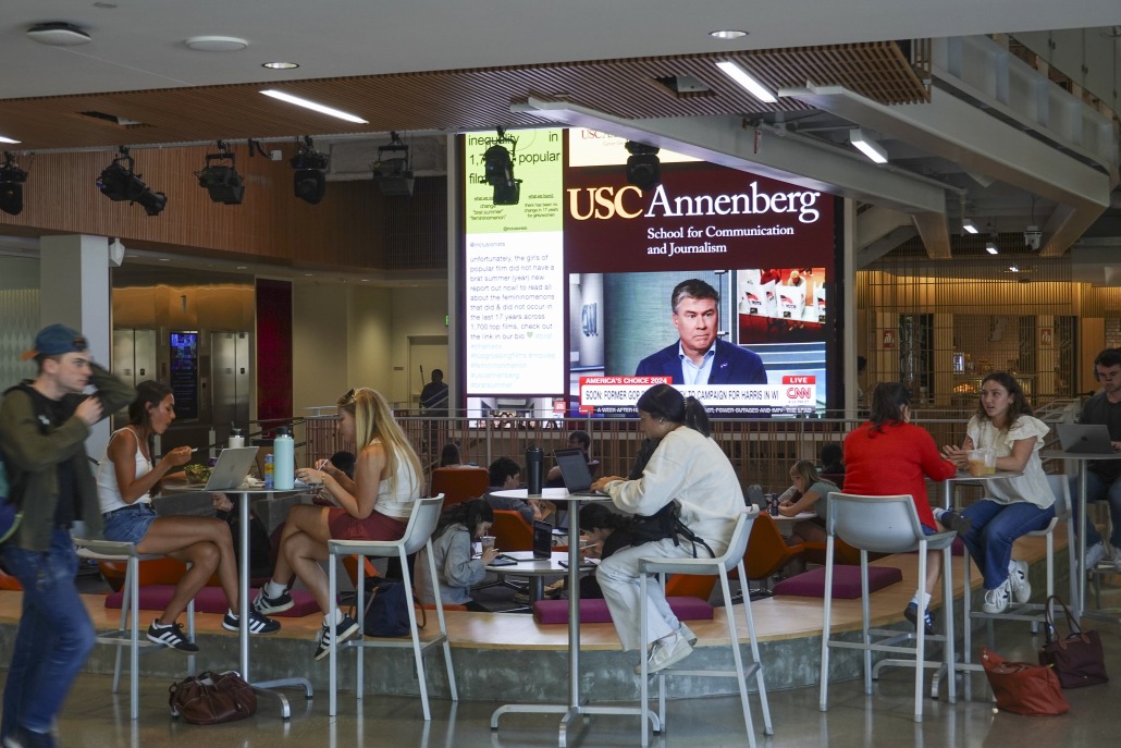 In the foreground, students sit at tables working on their computers. In the background, a big TV screen with the text "USC Annenberg School for Communication and Journalism" and live feed of a man in a blue suit speaking on CNN.