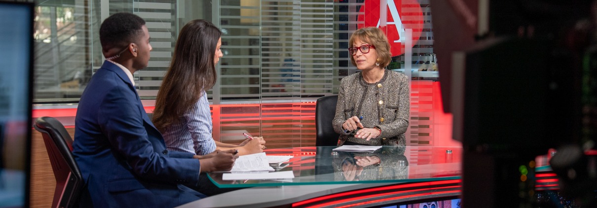 Carol Folt sits across from two student journalists. A camera is on the right side of the image.