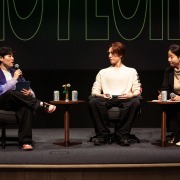 Three people sit in chairs onstage, with K-pop singer Yugyeom in the middle listening to a question from an interviewer to his right.