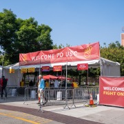 The front of the McCarthy Way entrance. A man wearing a blue jacket exits, and a sign above the tent containing the security scanners reads "Welcome to USC." To the right side of the tent, a sign reads, "Welcome to USC! Fight on!"