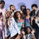 9 people pose for a group photo with a solid purple background behind them.