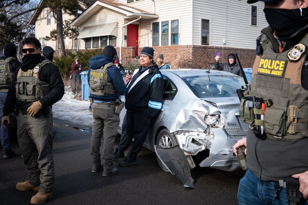 Immigration enforcement agents question man in front of a crashed car.