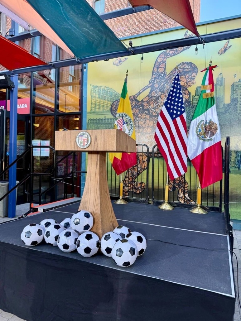 A podium stands before the L.A., U.S. and Mexican flags, with a pile of soccer balls.
