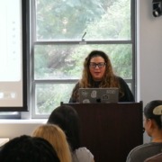 A professor stands at the front of a classroom in front of a projector screen