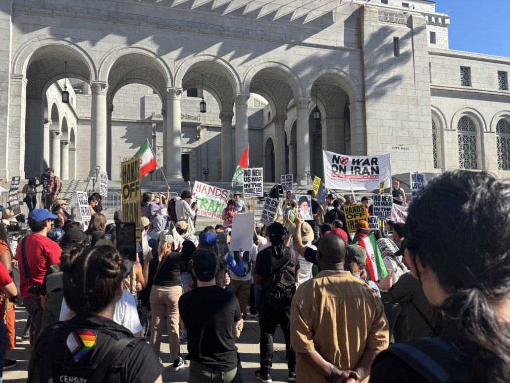 Protesters against the war in Iran hold signs and flags outside City Hall