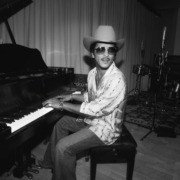 Bruno Mars wearing sunglasses and a cowboy hat sits at the piano.